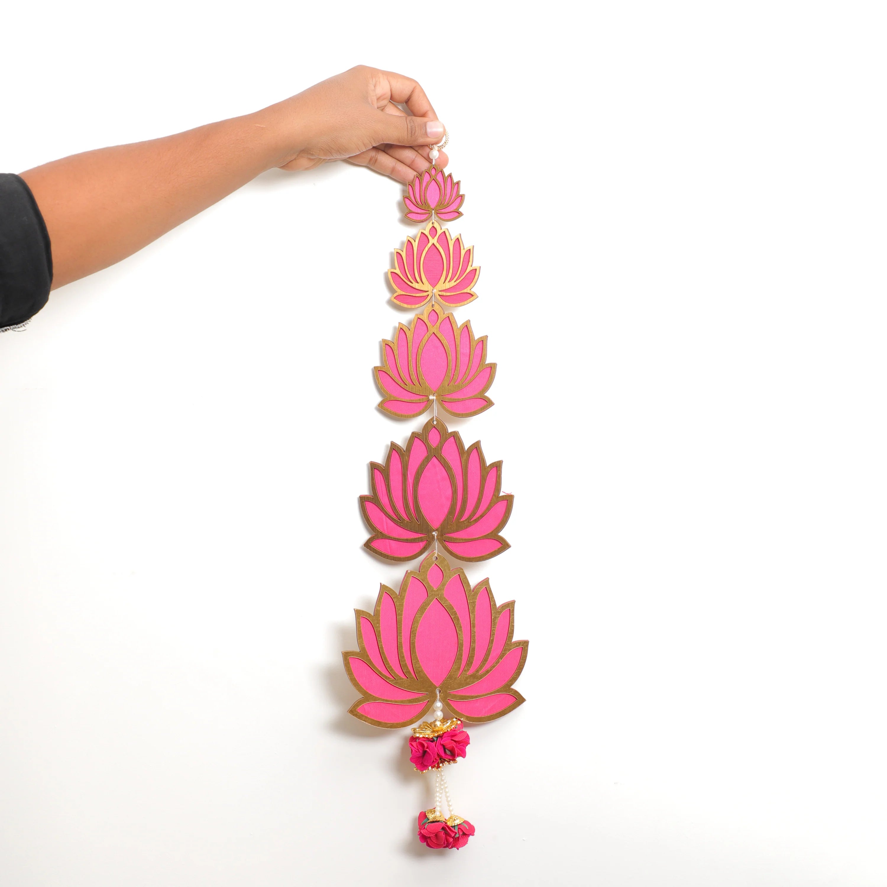 A hand holds a vertical decorative garland featuring five pink lotus flowers outlined in gold, with small pink tassels at the bottom, against a plain white background.
