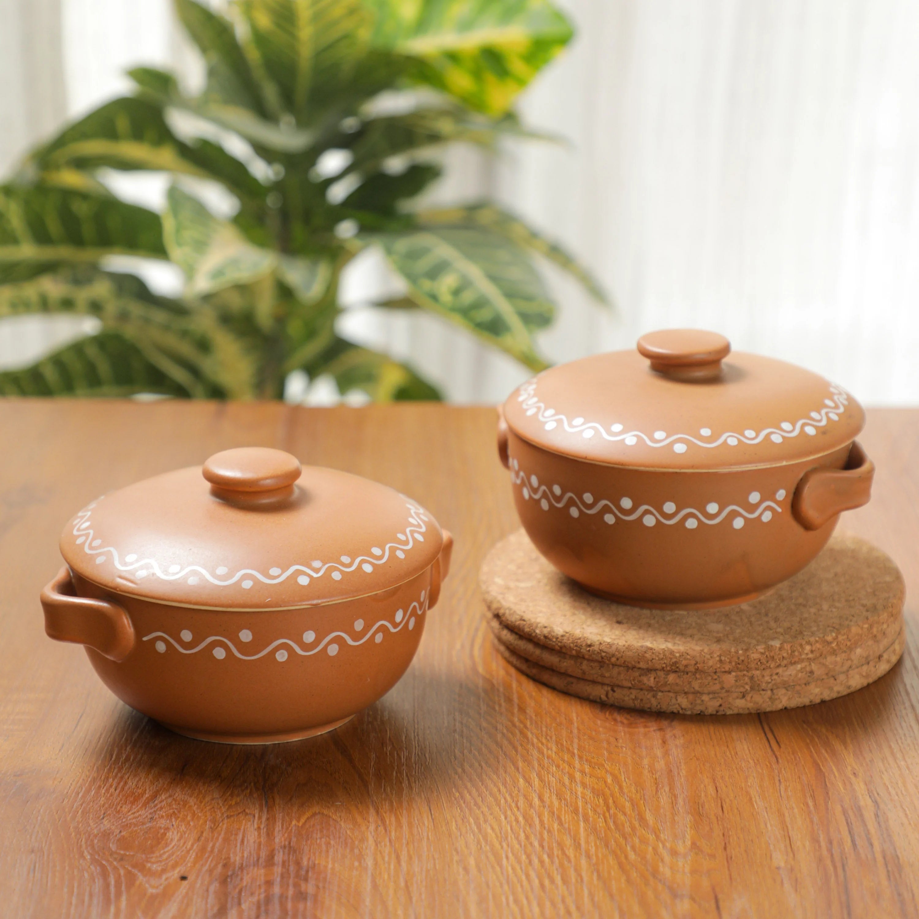 Two terracotta clay lidded pots with white decorative patterns, displayed on a wooden table with a blurred green plant in the background.
