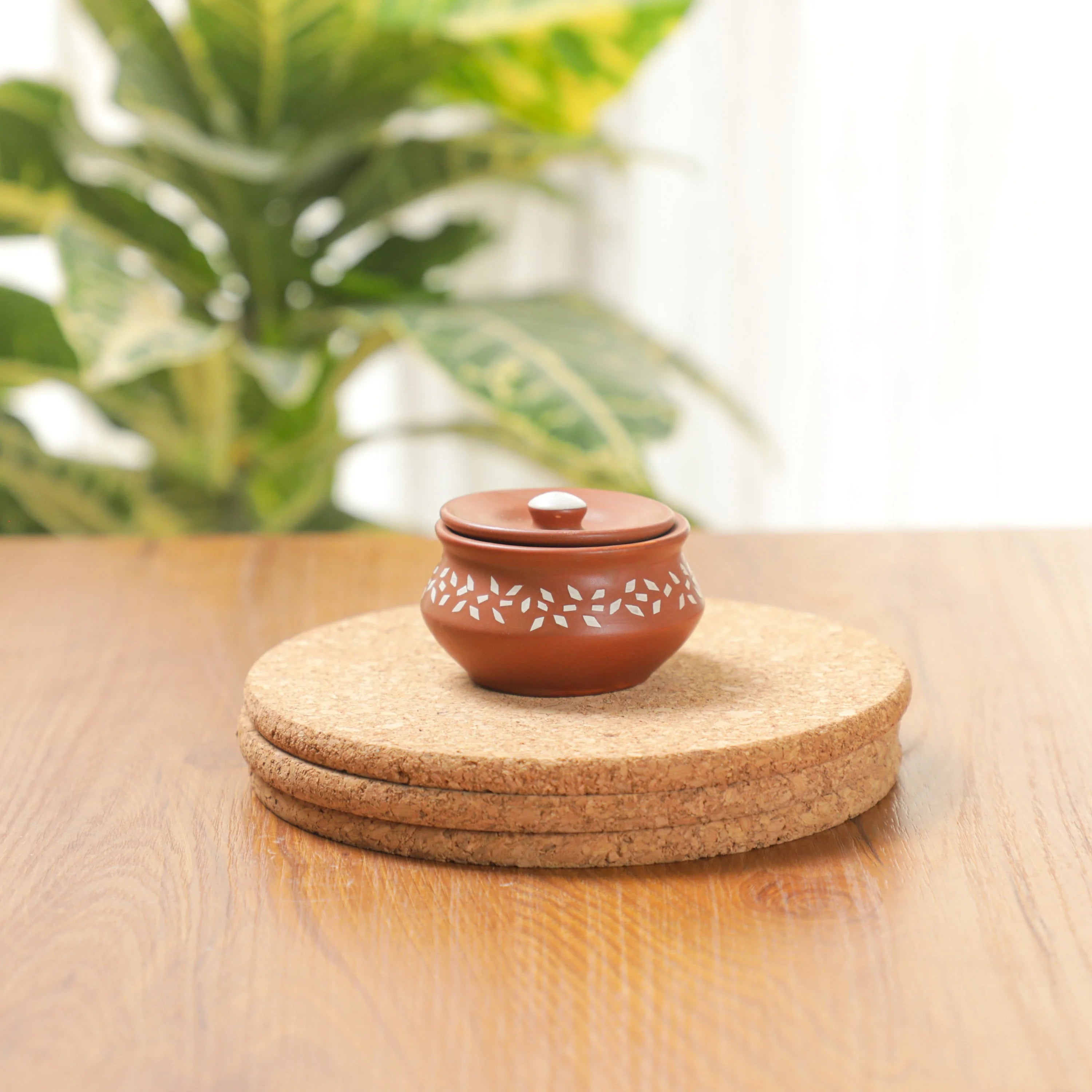 A small terracotta-colored ceramic lidded pot with white leaf patterns sits on a stack of round cork coasters on a wooden table, with a blurred green plant in the background.