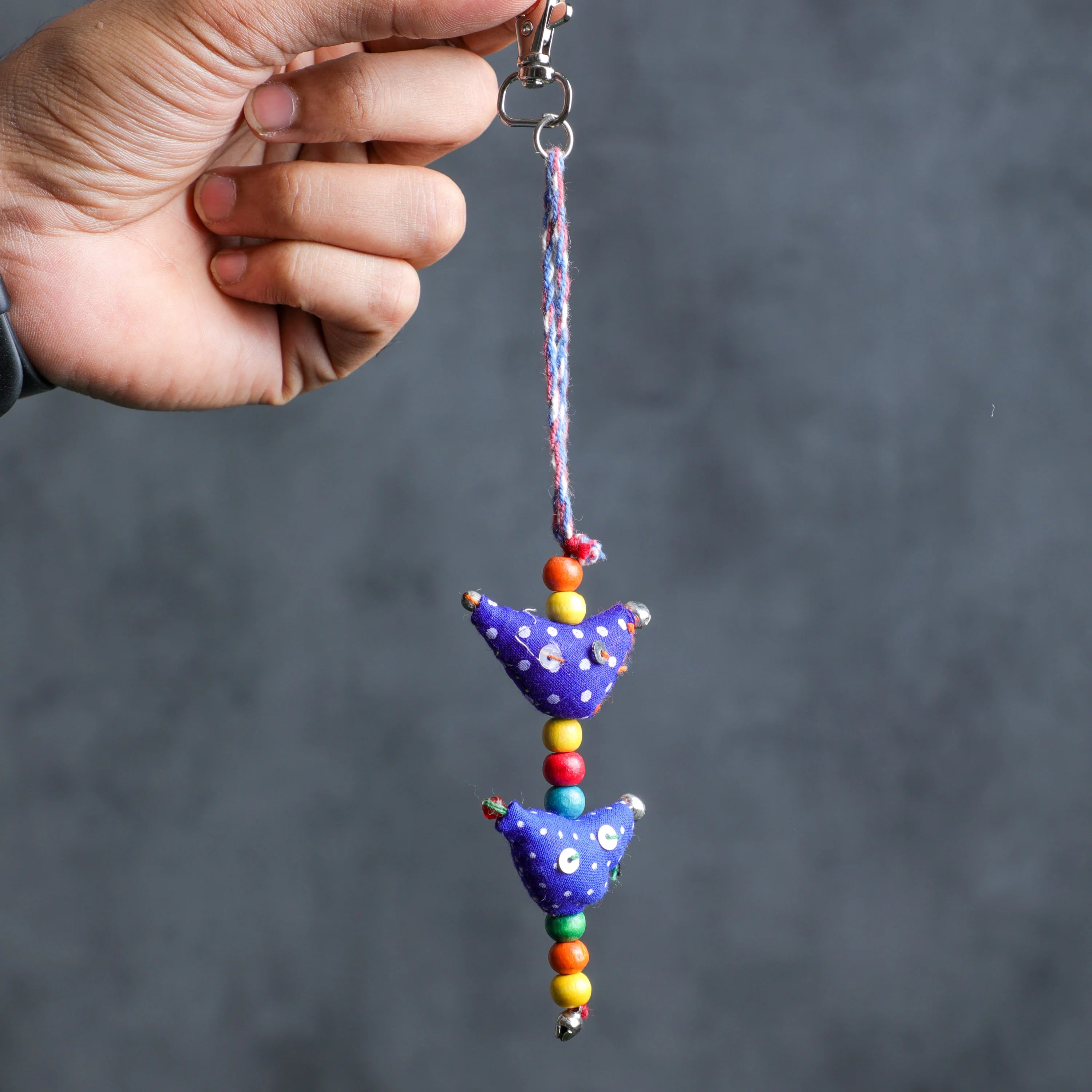 A hand holding a colorful fabric car keychain with two purple bird charms and multicolored beads, set against a dark gray background.