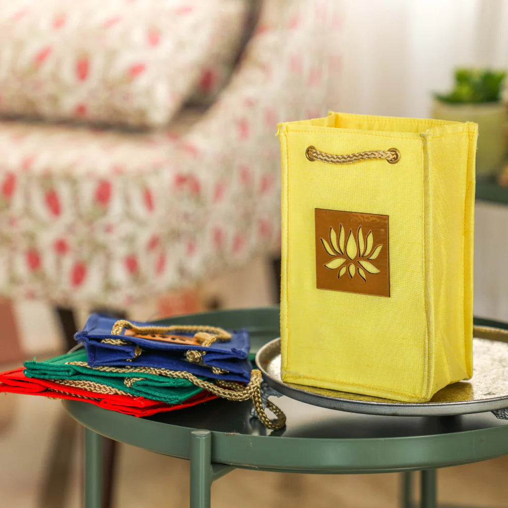A bright yellow fabric tote bag with a brown square patch featuring a gold lotus design, sitting on a round green table. A stack of folded colorful bags in red, green, and blue is visible on the left, with a blurred living room background.