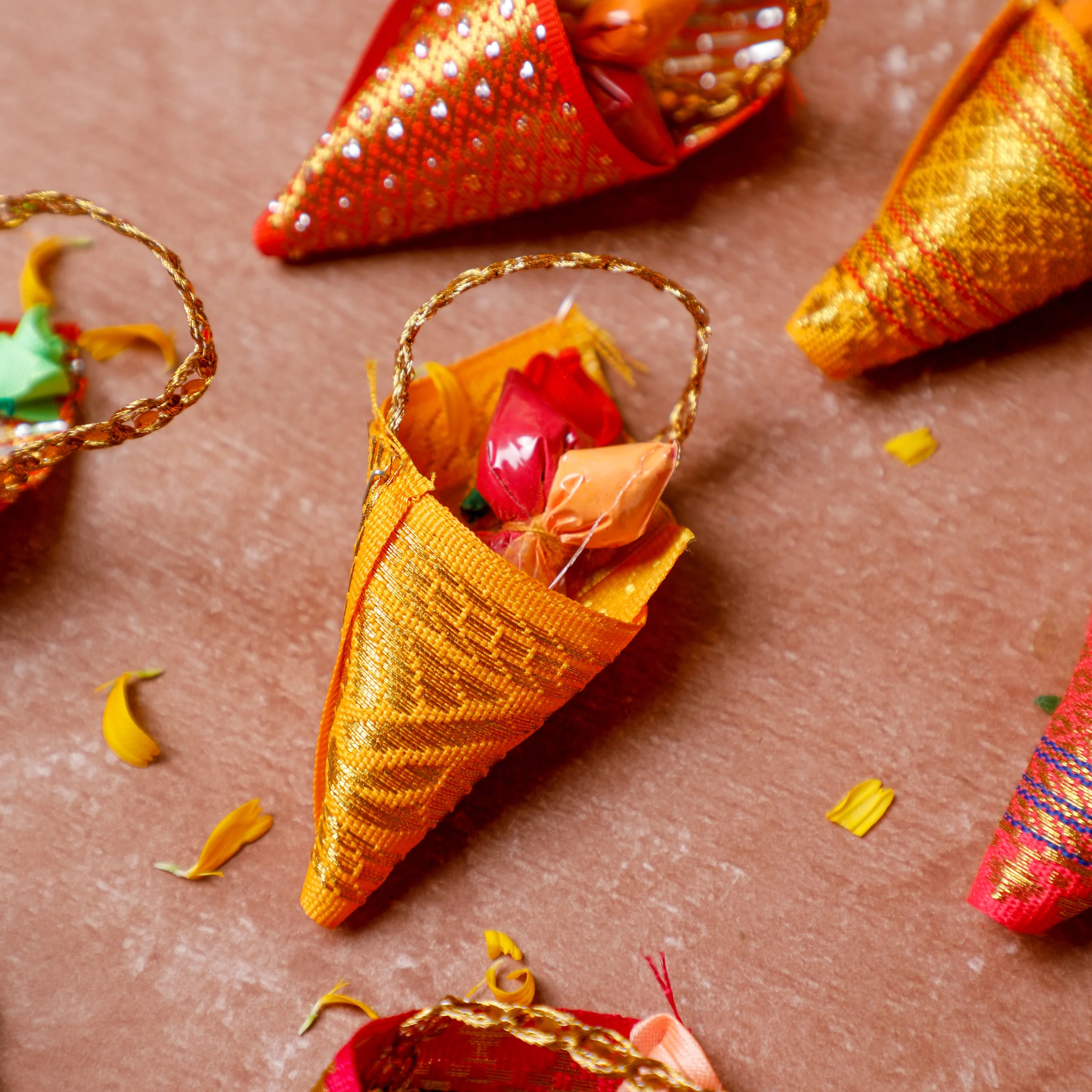 Decorative hand-woven fabric cone ornaments in orange, gold, and red, styled on a textured terracotta surface with scattered yellow flower petals and wrapped candies inside.