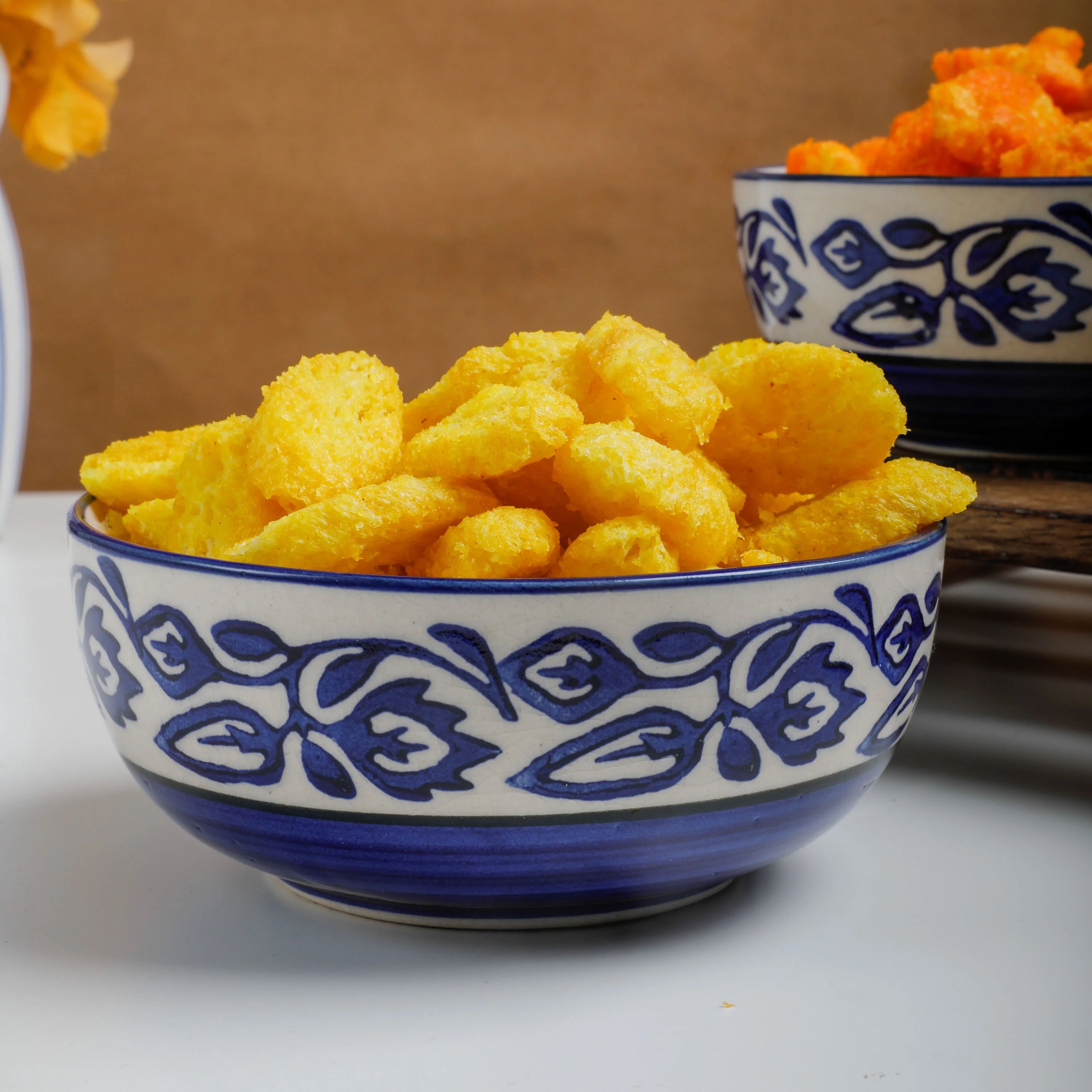 Yellow cheese-like snack pieces piled in a white ceramic bowl with a blue floral pattern, with a second matching bowl blurred in the background on a wooden surface.
