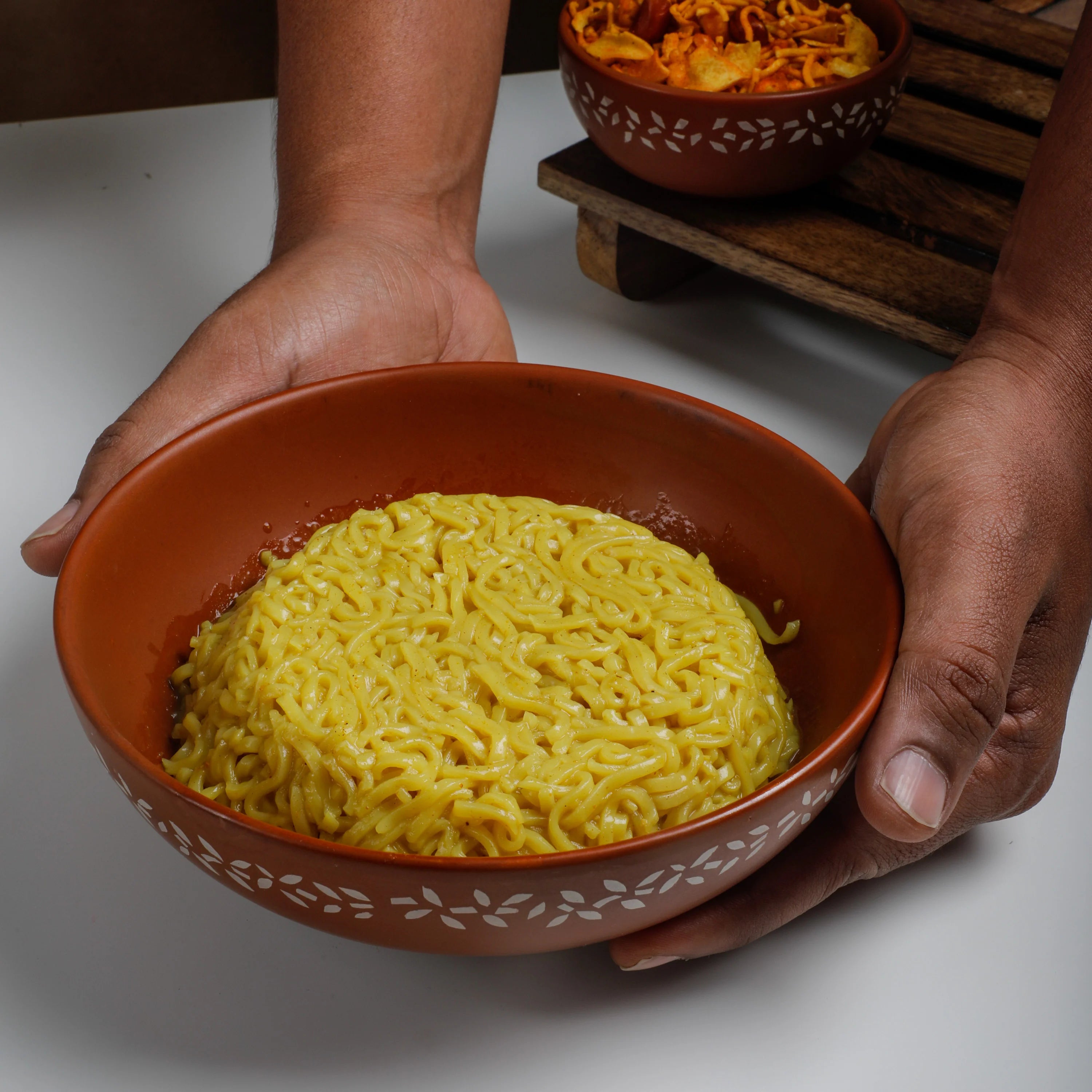 A person holds a terracotta-colored bowl filled with yellow instant noodles over a white surface, with another bowl in the background on a wooden stand.