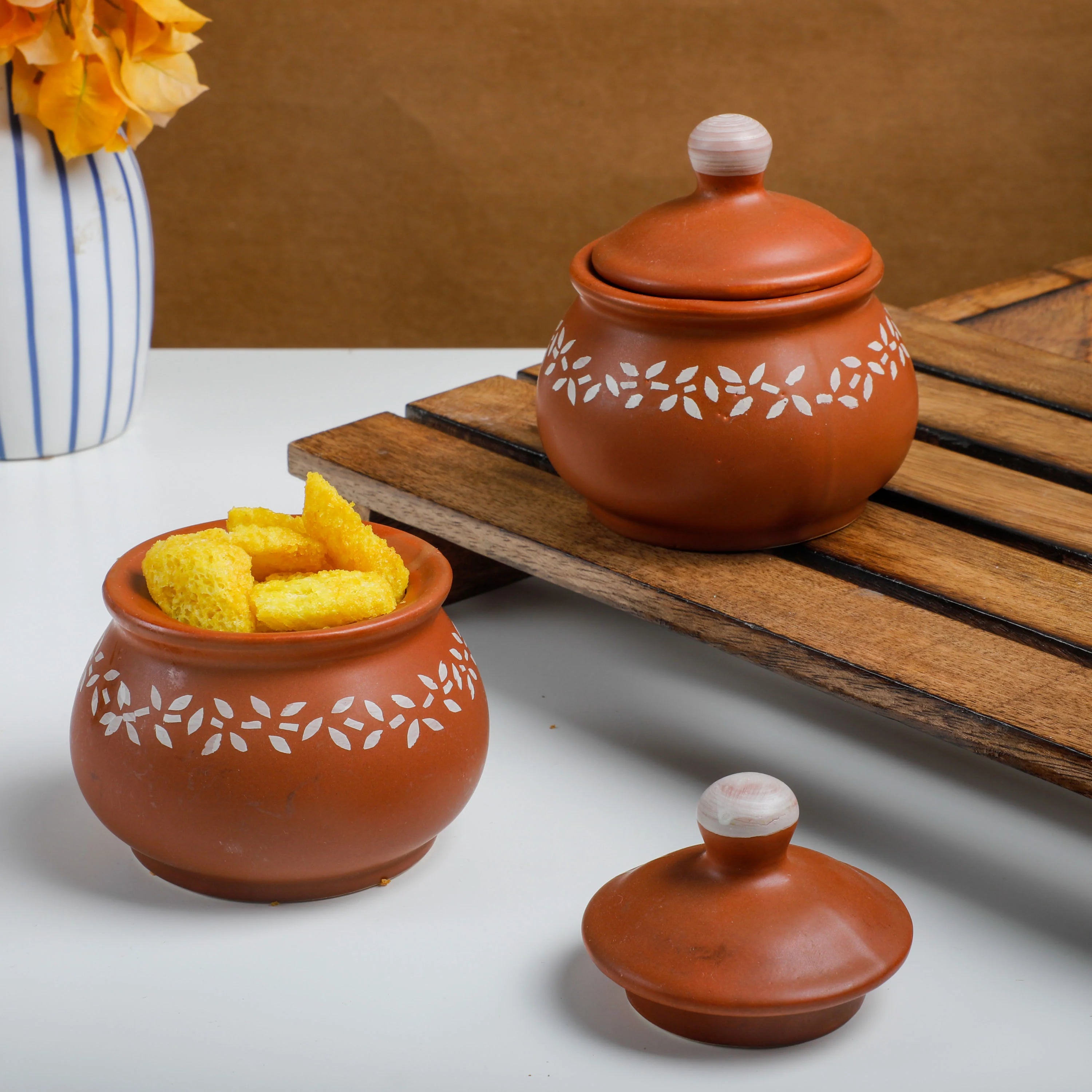 Two terracotta clay snack jars with white floral engravings, one filled with yellow corn snacks and the other closed on a wooden slatted tray, with a matching lid placed in front on a white surface.