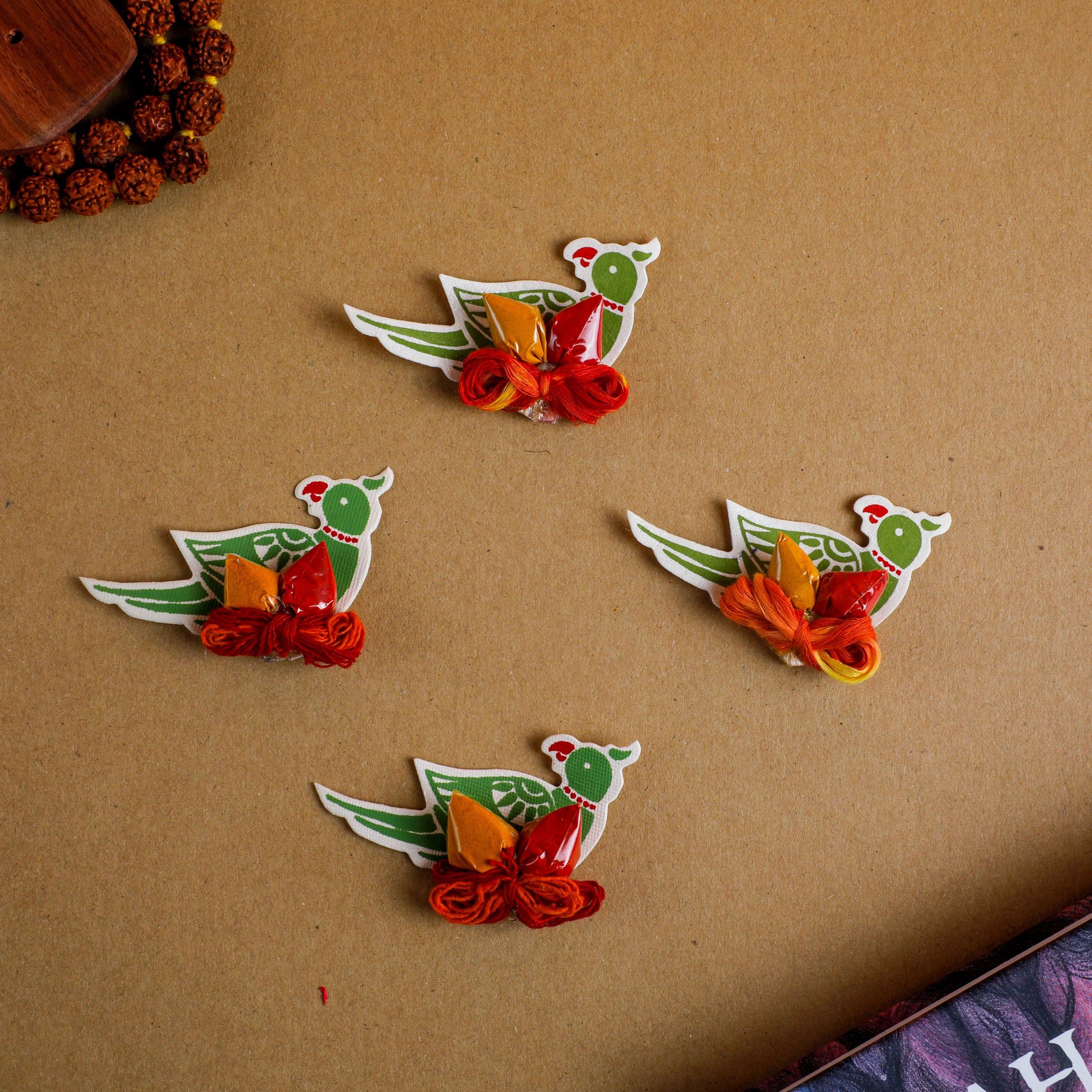 Four small decorative bird-shaped ornaments or charms arranged on a brown kraft-paper background. Each bird has a white outline, green body, red head detail, and red and orange wrapped ribbon accents.