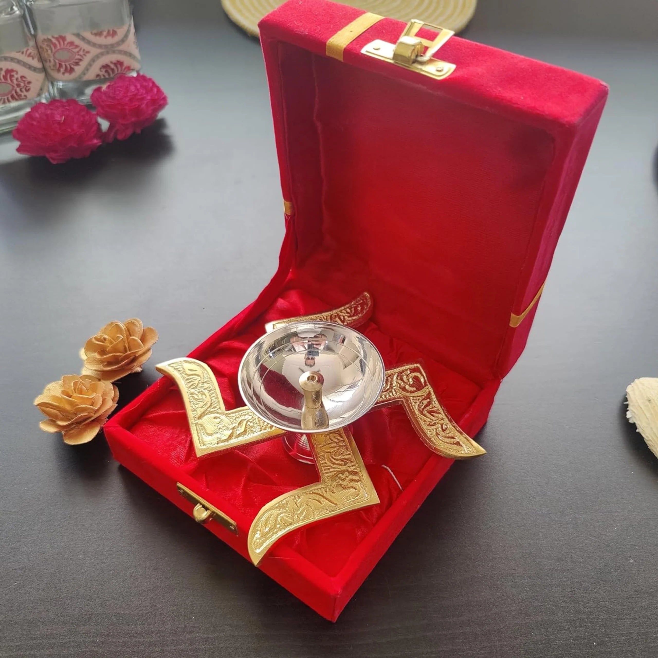 A clear glass prayer shawl (tallit) with white tassel, presented inside an open red velvet box with gold trim and decorative gold ribbons on a dark tabletop.